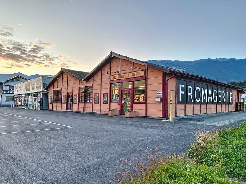 Large building, brown exterior, signage reading Fromagerie la Fromagerie, entrance door, windows, parked cars in the foreground, mountains in the background.