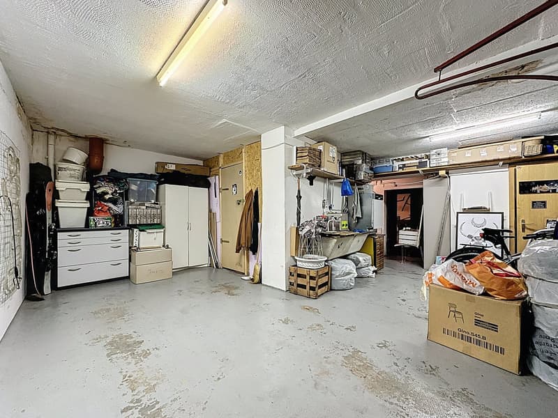 Empty room, concrete floor, white walls, large storage boxes, baskets, shelves with various items, door to another room, broom and cleaning products