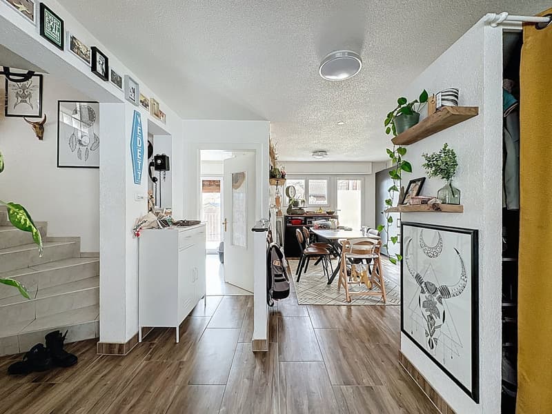 Spacious interior hallway with wooden floors, white walls, kitchen area with dining table, and framed pictures on walls.