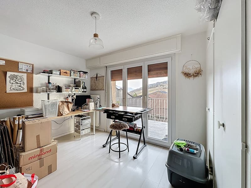 A room with a desk, a stool, boxes, a radiator, glass doors to a balcony, and mounted shelves