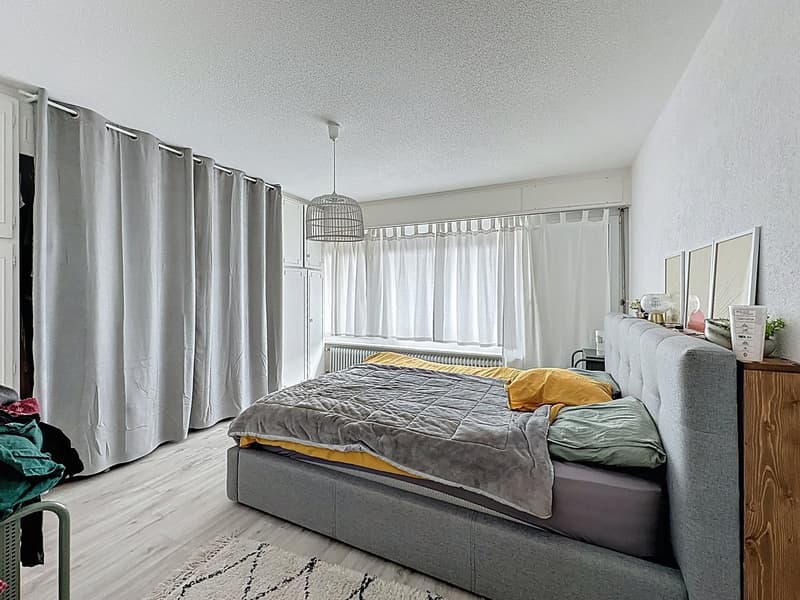Gray upholstered bed, white walls, wooden floor, white curtains, gray carpet, nightstand with plant