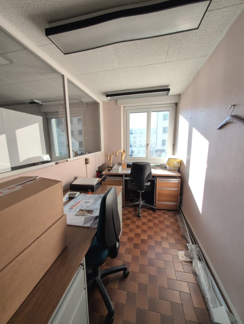 Office space with desks, chairs, boxes, and storage units. One desk has a large mirror on the wall behind it. Large window on the wall opposite of the desk.