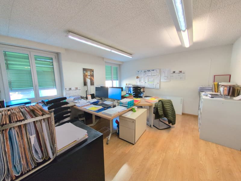 Office with wooden floor, white walls, two desks, monitors, chairs, cabinets, papers, and ceiling lights