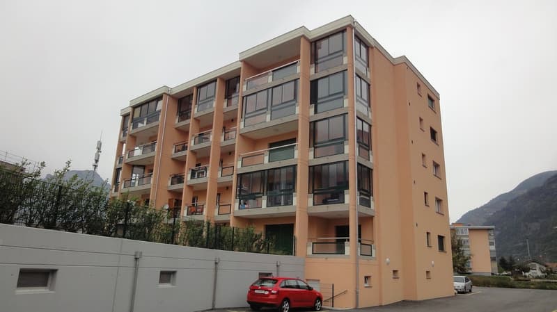 Multi-story apartment building with balconies, beige exterior, multiple windows, red car parked in front