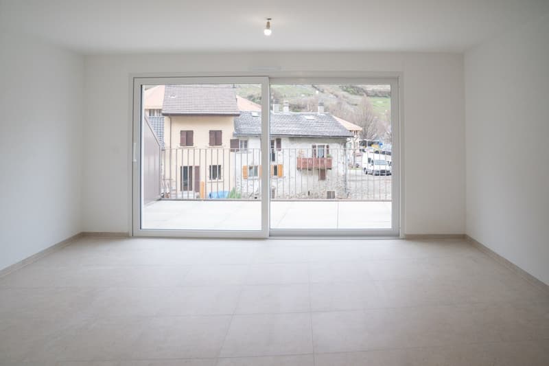 Empty room with tiled floor, large sliding glass door leading to a balcony, white walls, ceiling lamp, distant view of houses and a mountain