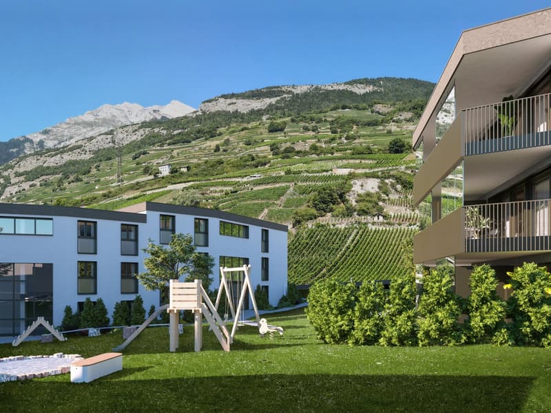 A two-story white building with a sloped roof, balconies, and windows. There is a terrace with a swing set and slide in front. The building is surrounded by green grass and bushes.