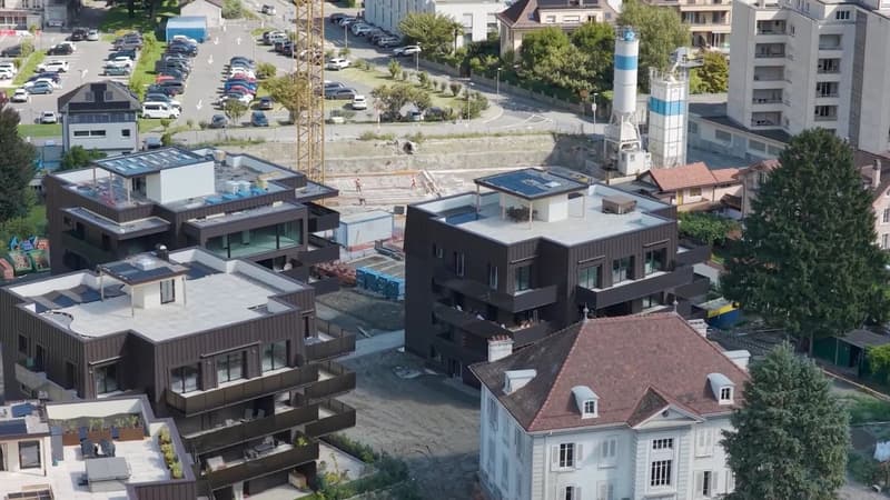 Multiple residential buildings under construction, with modern architecture and several cars parked in a parking lot. Each building features a flat roof with solar panels, suggesting an emphasis on sustainability. One of the buildings shows a swimming poo