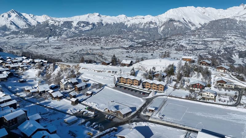 Aerial view of a residential area with multiple chalet-style houses covered in snow. In the background are mountains also covered in snow.