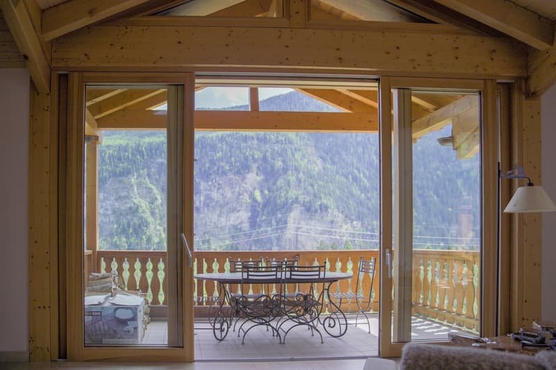 Wooden cabin interior with large windows overlooking a forested mountain landscape, a round wooden dining table with wrought iron chairs on a balcony or terrace