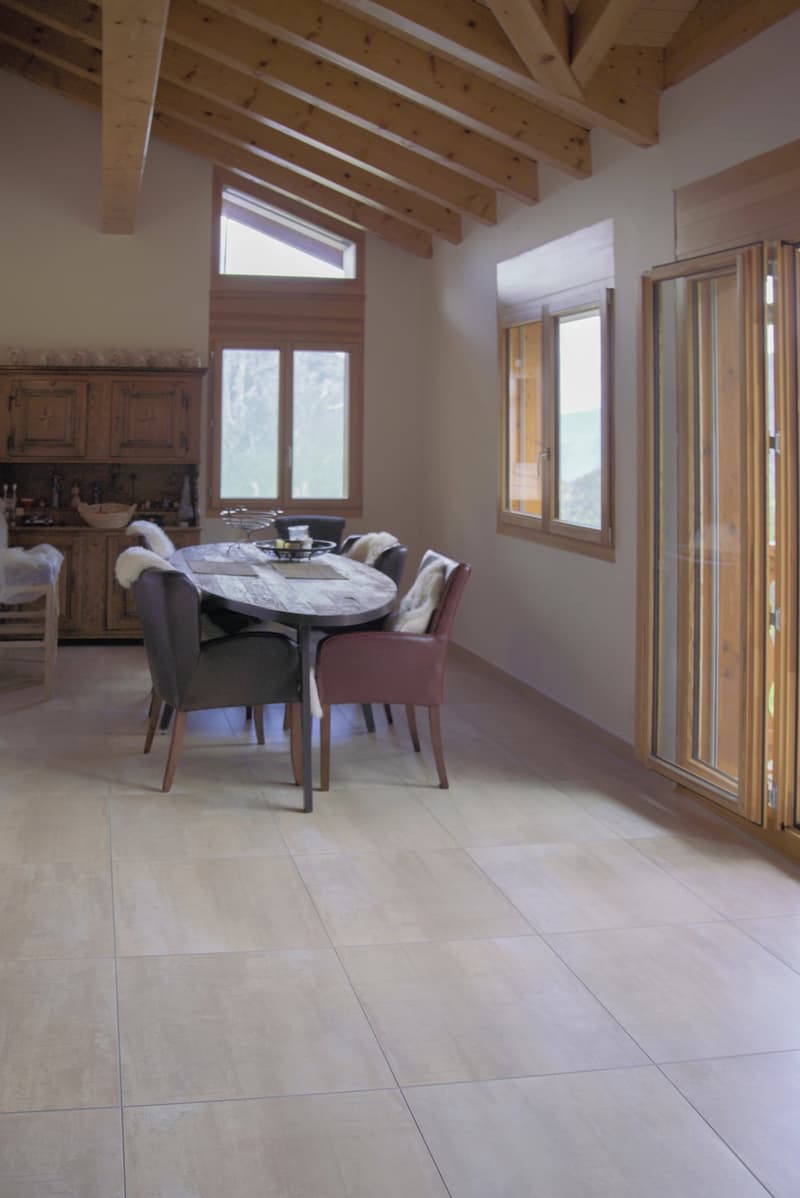 Spacious living room with wooden beamed ceiling, large windows, rustic wooden dining table and chairs, and tile flooring.