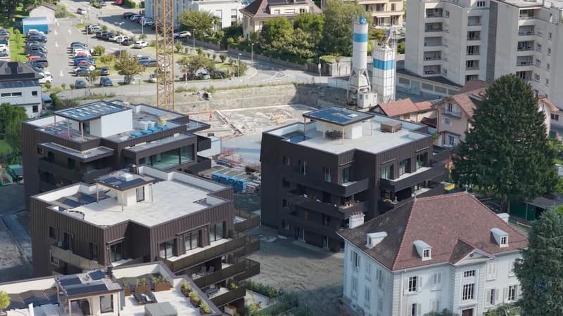 Two newly built contemporary houses with terraces, surrounded by an area under construction and other residential buildings, with cars parked in the parking lot