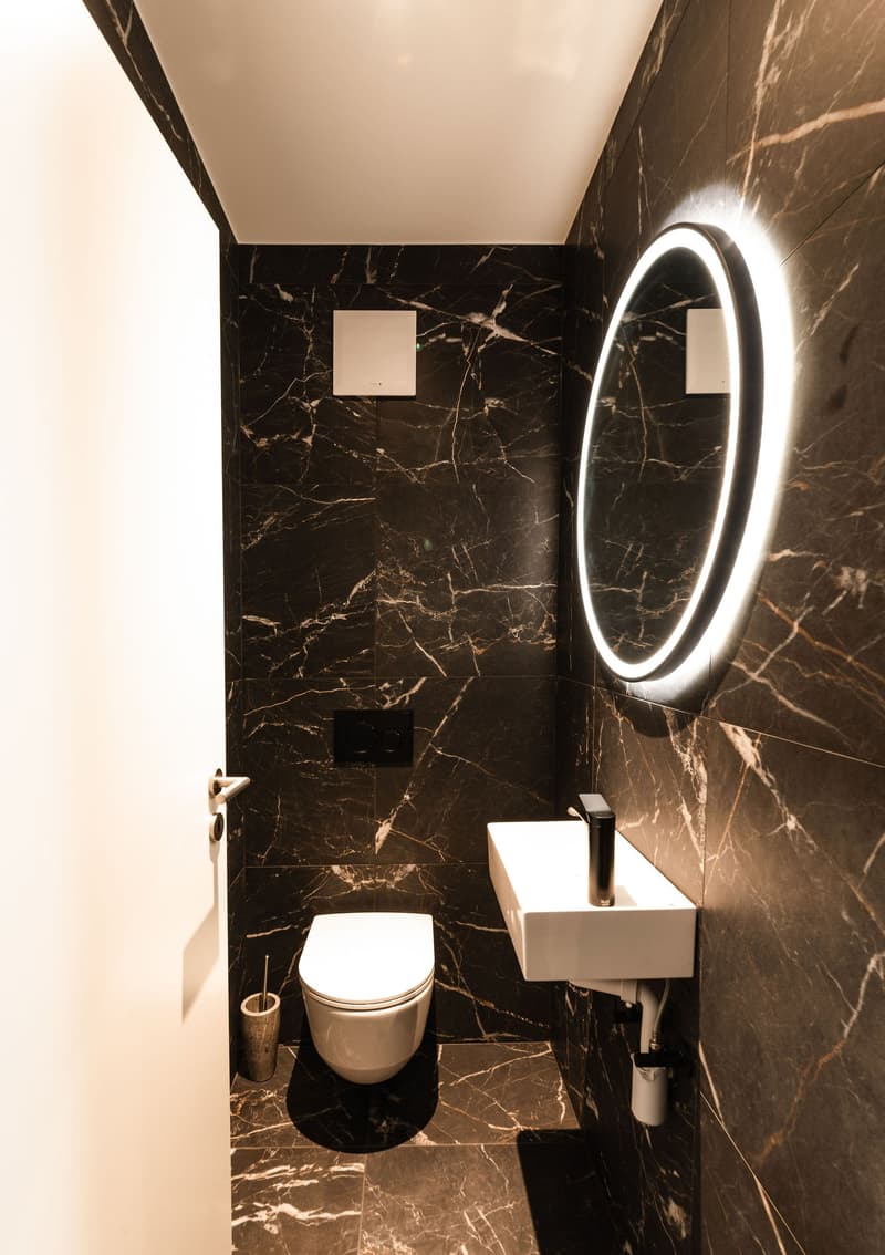 small bathroom with black marble walls, white toilet, white sink, round mirror with light