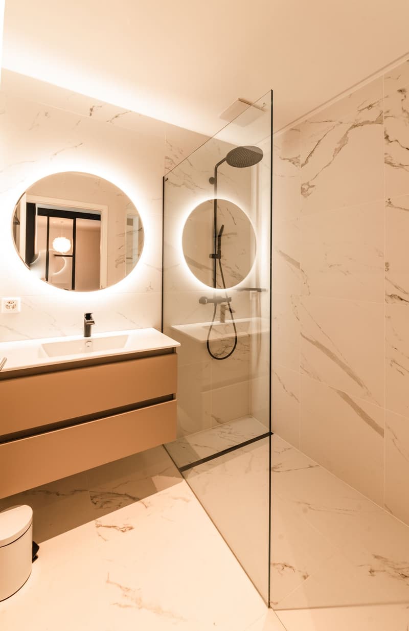 modern bathroom with white marble tiles, glass shower enclosure, round mirrors, wooden vanity with sink, and a toilet
