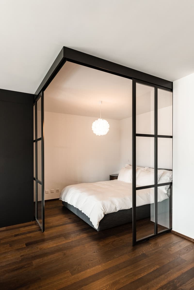 bedroom with glass walls, wooden floor, white bed, white pillows, white ceiling, white walls, black ceiling trim, white light fixture