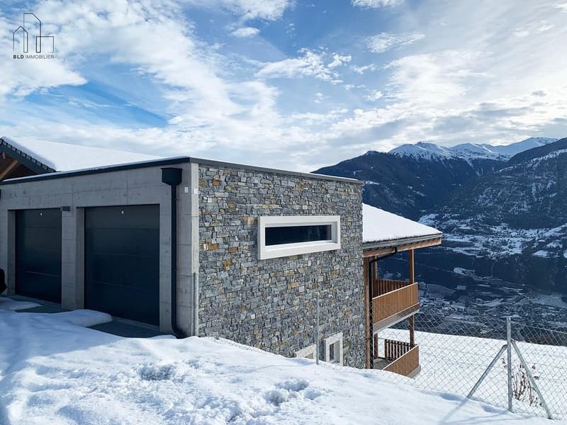 Modern house with stone facade, two garages, wooden balcony, snowy roof, mountainous background
