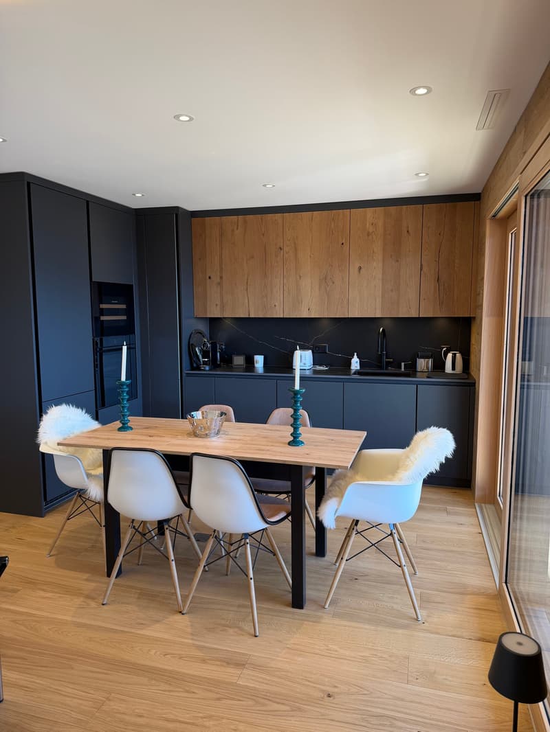 Dining room with wooden table, white chairs, black cabinets, wooden floor, glass door, kitchen area with sink, countertop, and appliances