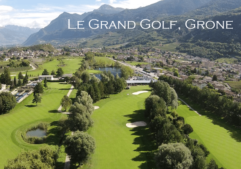 Aerial view of a large, well-maintained golf course surrounded by mountains, forests, and a small town. The course features several fairways, sand traps, and a lake or pond. There appears to be a parking area near the clubhouse or facilities.