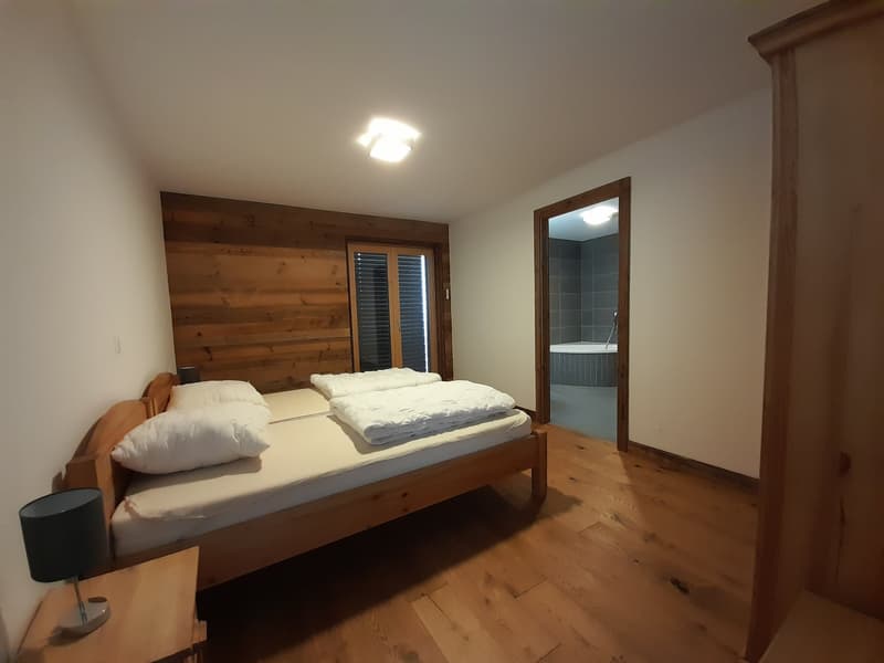 Wooden bed, two pillows, wooden cabinet, wall lamp, bathroom with tub, wooden wall