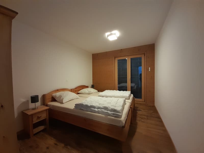 small bedroom with twin beds, white walls, wooden floor, nightstand with lamp, small windows