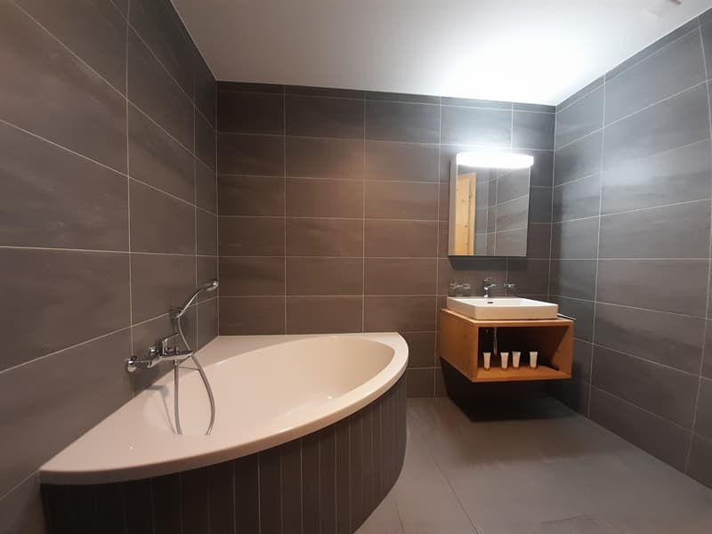 white bathtub, grey tiled walls, wooden vanity, sink, mirror