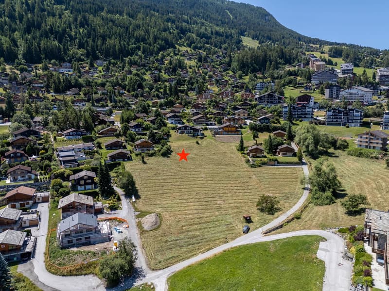 Plot of land in a mountainous area with surrounding houses, a wide green field, and a mountain in the background