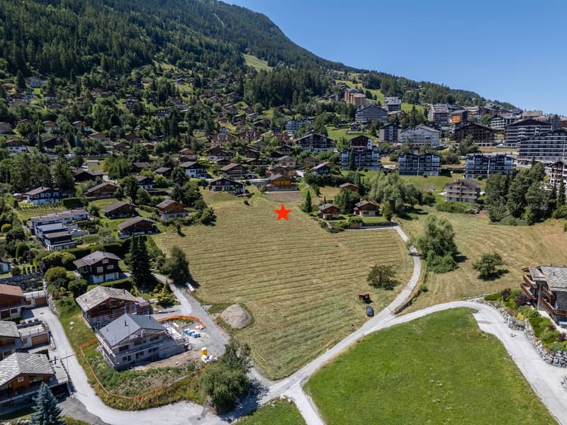 Plot of land, wooded surroundings, several buildings around, mountainous landscape, open field in the middle