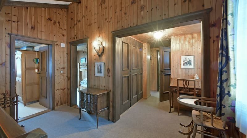 A corridor with wooden walls, two beds, a desk and chairs, a cabinet with a lamp on the wall, a picture on the wall, a desk, a chair, and a shelf with pillows and a picture frame.