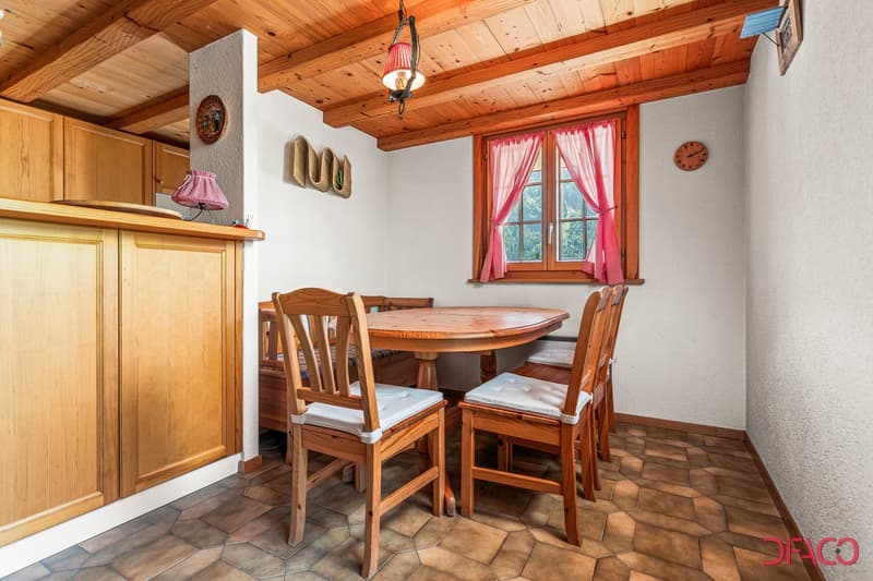 Cozy wooden dining room with a round wooden table, wooden chairs, and a fireplace. The room has a rustic, cabin-like feel with wooden ceiling beams and walls. There are pink curtains on the window and decorative wall accents.