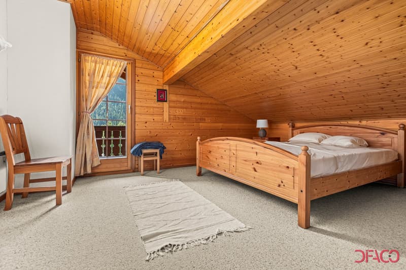 Spacious bedroom with wooden paneled walls and ceiling, large window with curtains, wooden bed frame, wooden chair, and a rug on the floor.