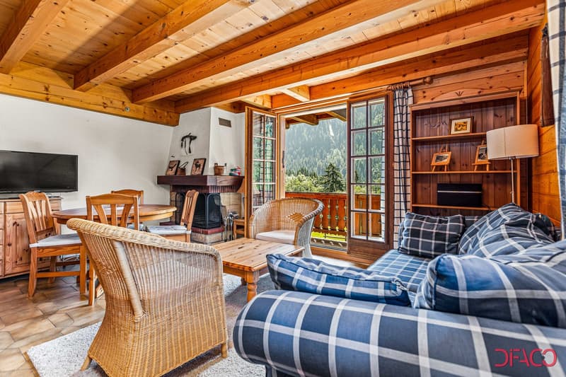 Cozy wooden cabin interior with exposed beams, large windows overlooking a scenic mountain view, a fireplace, wicker furniture, and a dining area with a wooden table and chairs.