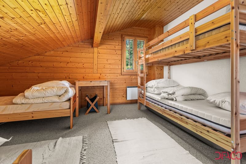Cozy wooden cabin bedroom with bunk beds, a single bed, and a small side table. The room has a warm, rustic feel with the wooden walls and ceiling.