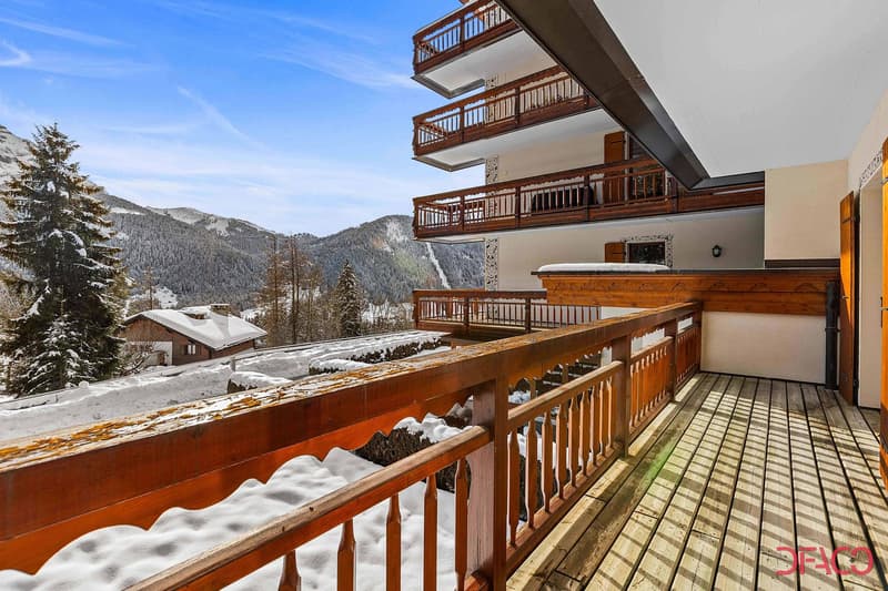 Multi-story wooden building with balconies overlooking a snowy mountain landscape. The balconies have wooden railings and the building appears to be in a ski resort or alpine setting.