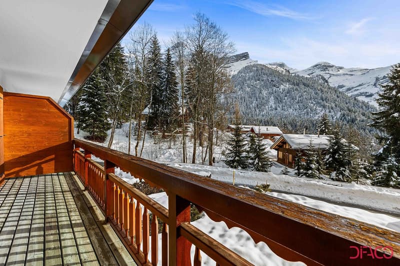 Wooden balcony overlooking a snowy mountain landscape with pine trees and chalets in the distance