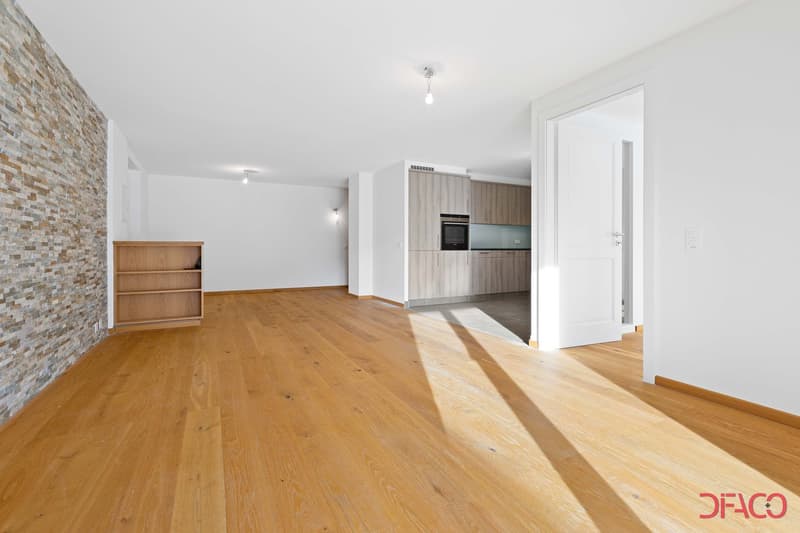 Spacious open-concept living area with hardwood floors, a stone accent wall, and a modern kitchen with built-in appliances including a dishwasher. The room has ample natural light and a clean, minimalist design.