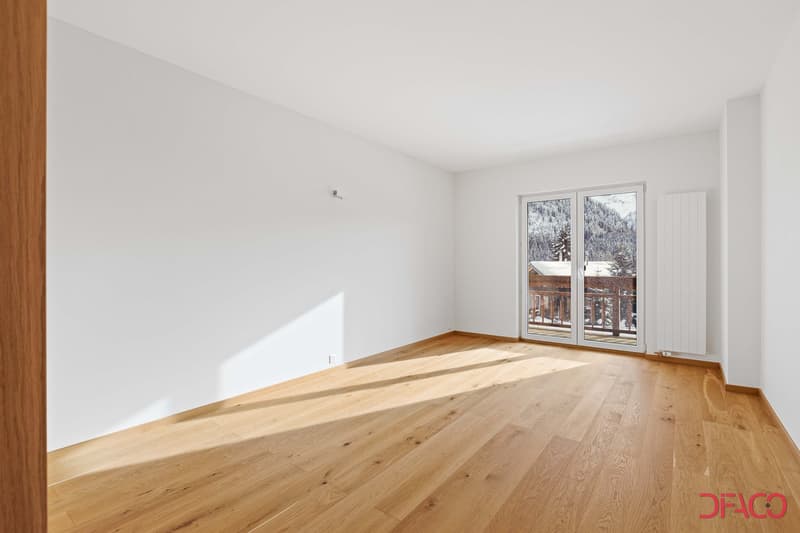 Spacious room with hardwood flooring, white walls, and large windows overlooking a snowy outdoor scene. The room appears to have access to a balcony or terrace.
