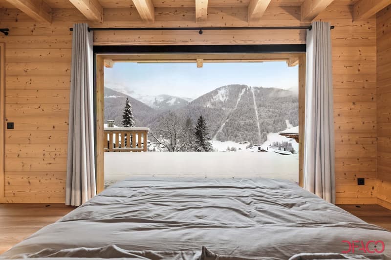 Cozy wooden bedroom with large windows overlooking a snowy mountain landscape. The room features a comfortable bed, wooden beams on the ceiling, and a balcony or terrace with a scenic view of the snow-covered mountains and trees.