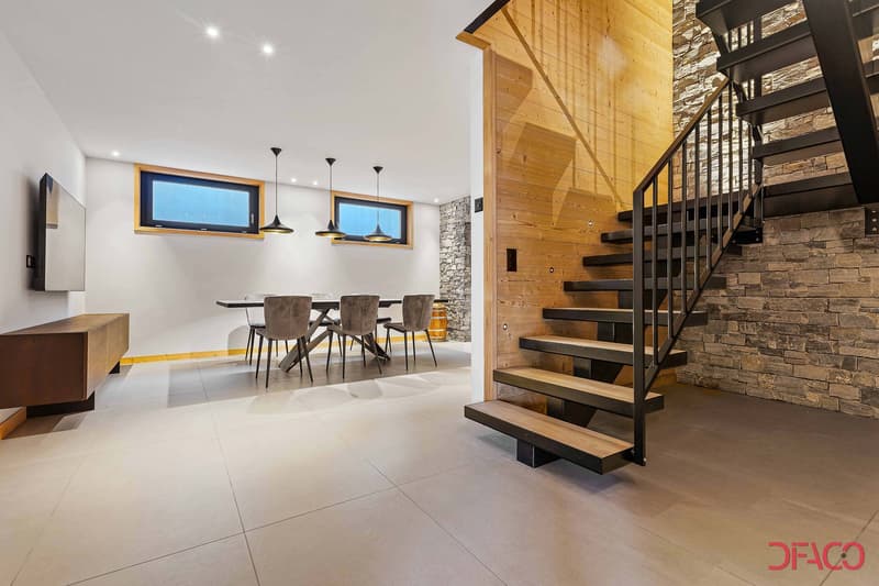 Open-concept living and dining area with wooden beams, stone accent wall, modern lighting fixtures, large windows, and a floating staircase with metal railings.