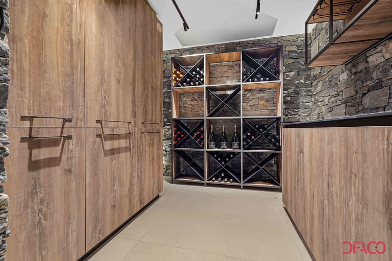 The image shows a wine cellar with a large wooden wine rack that takes up the entire wall. The wine rack has multiple compartments for storing wine bottles. The walls are made of stone, giving the space a rustic and cozy feel. There is also a fireplace vi
