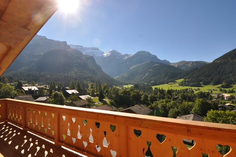 Wooden balcony with heart-shaped cutouts, overlooking a scenic mountain landscape with forests, villages, and snow-capped peaks in the distance under a bright, sunny sky