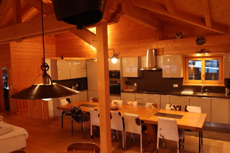 Spacious open-plan kitchen and dining area with wooden beamed ceiling, large windows, wooden dining table and chairs, kitchen cabinets and appliances, and a fireplace visible in the background.
