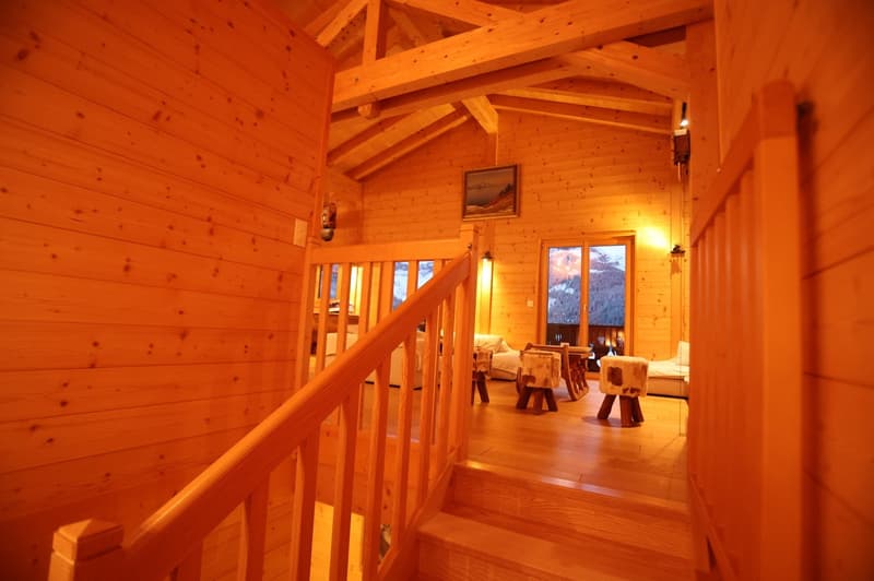 Cozy wooden cabin interior with exposed beams, wooden walls and floors, a fireplace, and rustic furniture such as stools and a table. Large windows provide a scenic view of the snowy landscape outside.