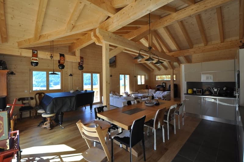 Spacious open-plan living area with high wooden ceilings and beams, large windows providing scenic mountain views, dining table, living room furniture, and a modern kitchen with stainless steel appliances.