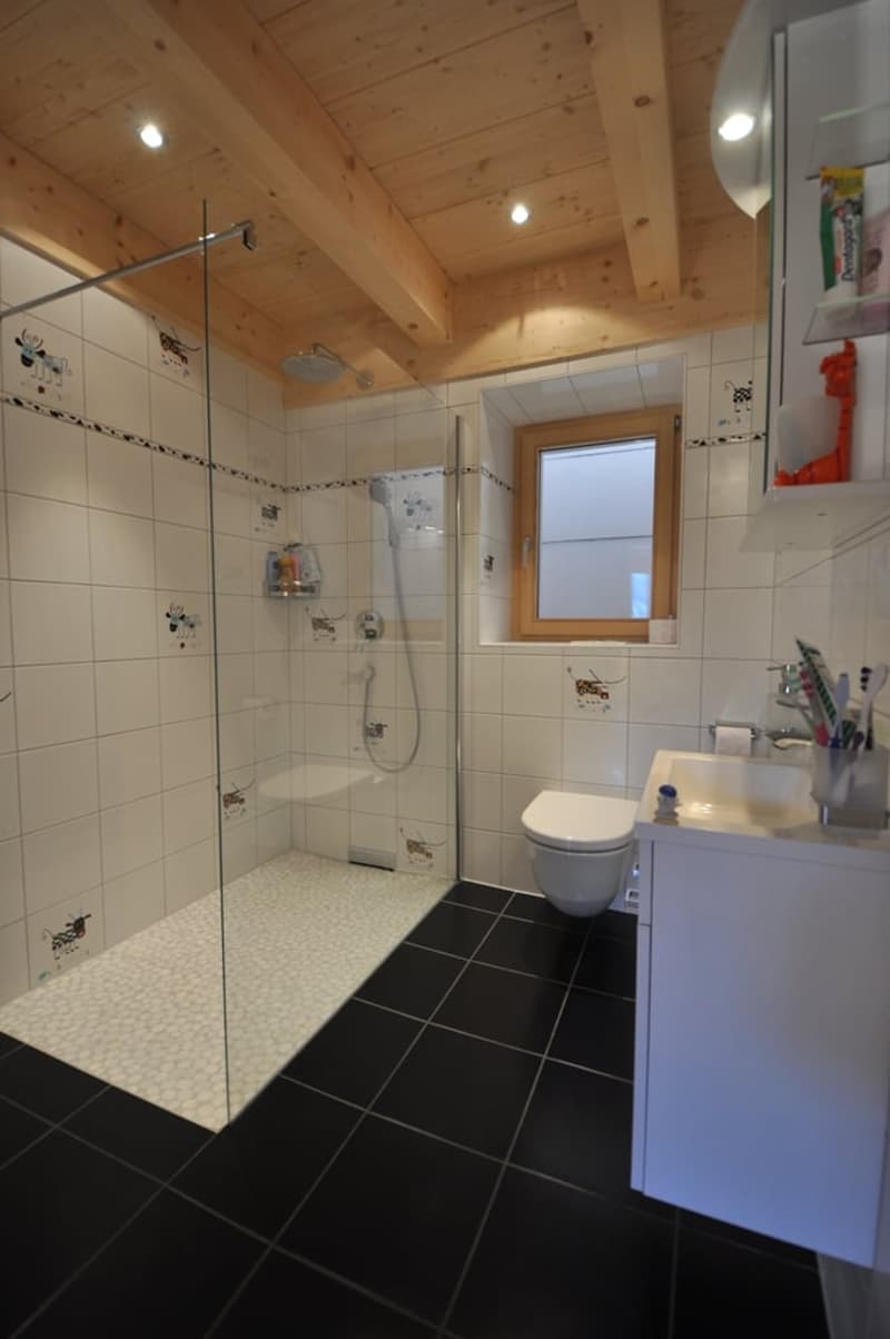 Spacious bathroom with wooden ceiling, tiled walls and floor, glass shower enclosure, vanity unit with sink, and a window