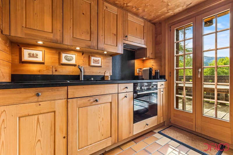 Rustic wooden kitchen with built-in cabinets, black countertops, stainless steel appliances including a dishwasher, and large windows overlooking a scenic outdoor view.