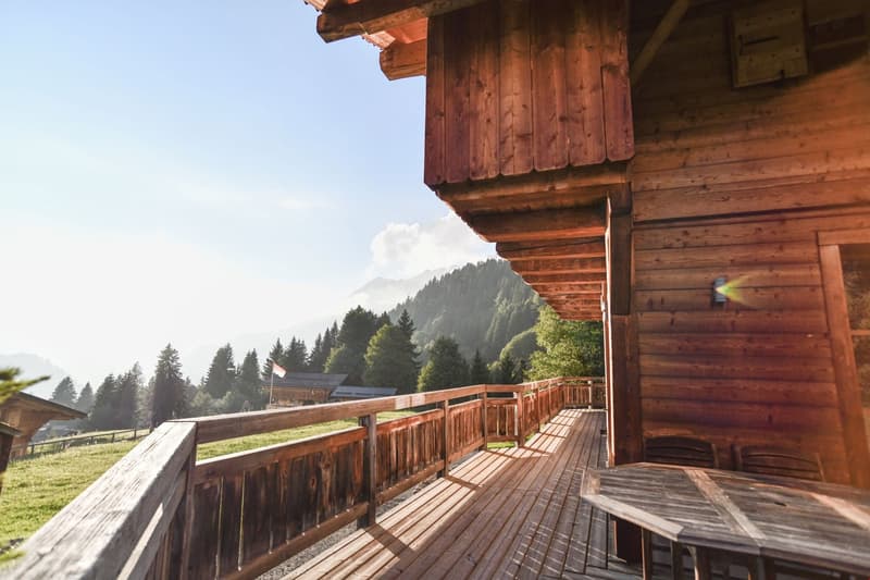 Wooden cabin with a large balcony or terrace overlooking a forested mountain landscape. The cabin has a rustic, traditional design with wooden walls and a slanted roof. The balcony provides a scenic view of the surrounding nature.