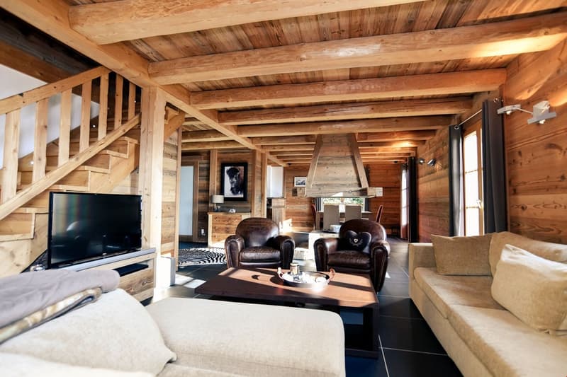 Rustic wooden cabin interior with exposed beams, leather armchairs, coffee table, and a large flat-screen TV mounted on the wall