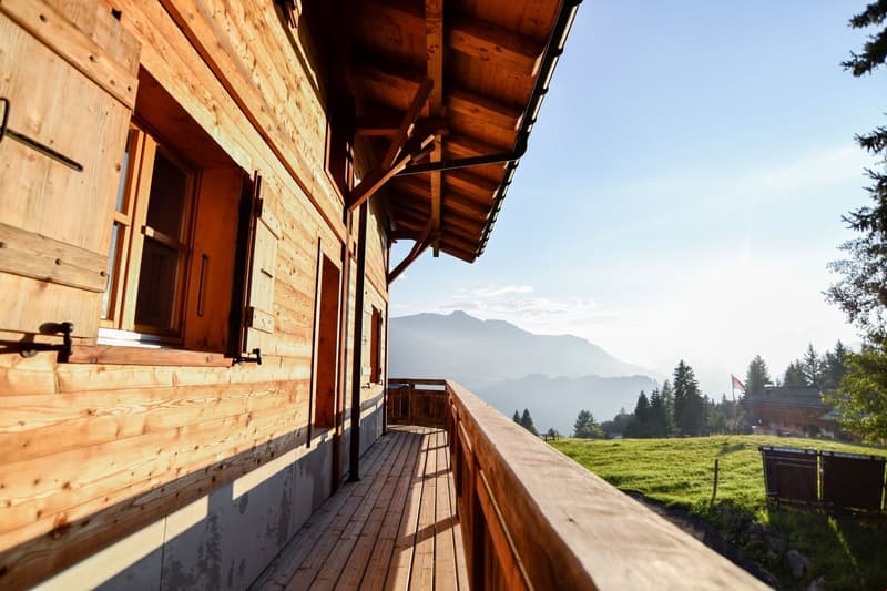 Wooden cabin with a covered balcony/terrace, surrounded by a grassy field and mountains in the background.