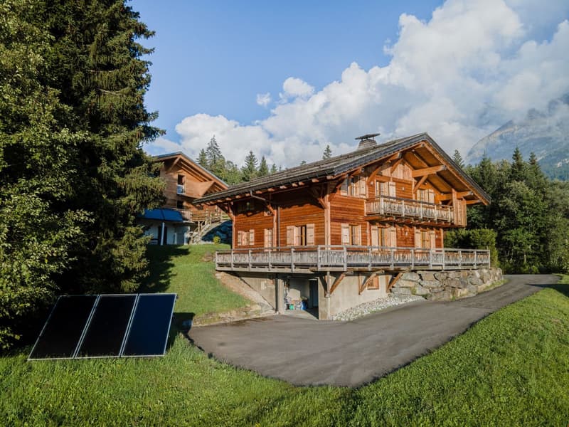 Two-story wooden chalet-style house with balconies, surrounded by trees and mountains, with a paved driveway and solar panels visible.
