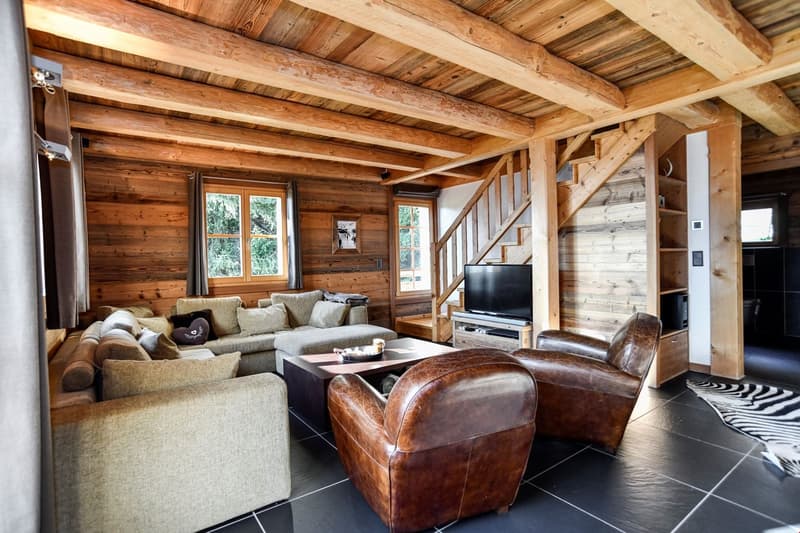 Rustic wooden cabin interior with exposed beams, large windows, comfortable sofas and armchairs, a coffee table, and a TV stand. The room has a cozy and warm atmosphere.