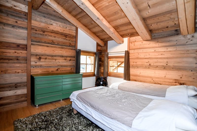 Rustic wooden cabin bedroom with exposed wood beams and paneling, a large bed with white bedding, a green dresser, and a fireplace visible in the corner.
